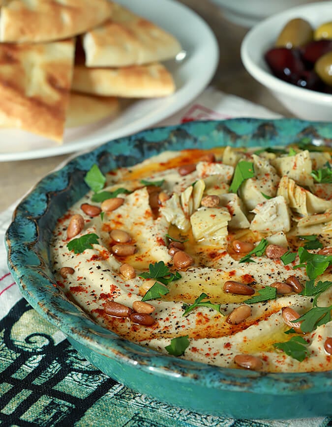 Artichoke and Roasted Garlic Hummus in a bowl is garnished with chopped artichoke, pine nuts, and parsley.