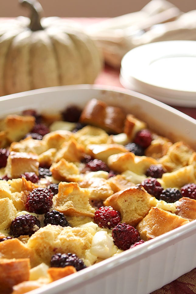 A white dish is filled with bread, beaten eggs, and blackberries for a baked dish.