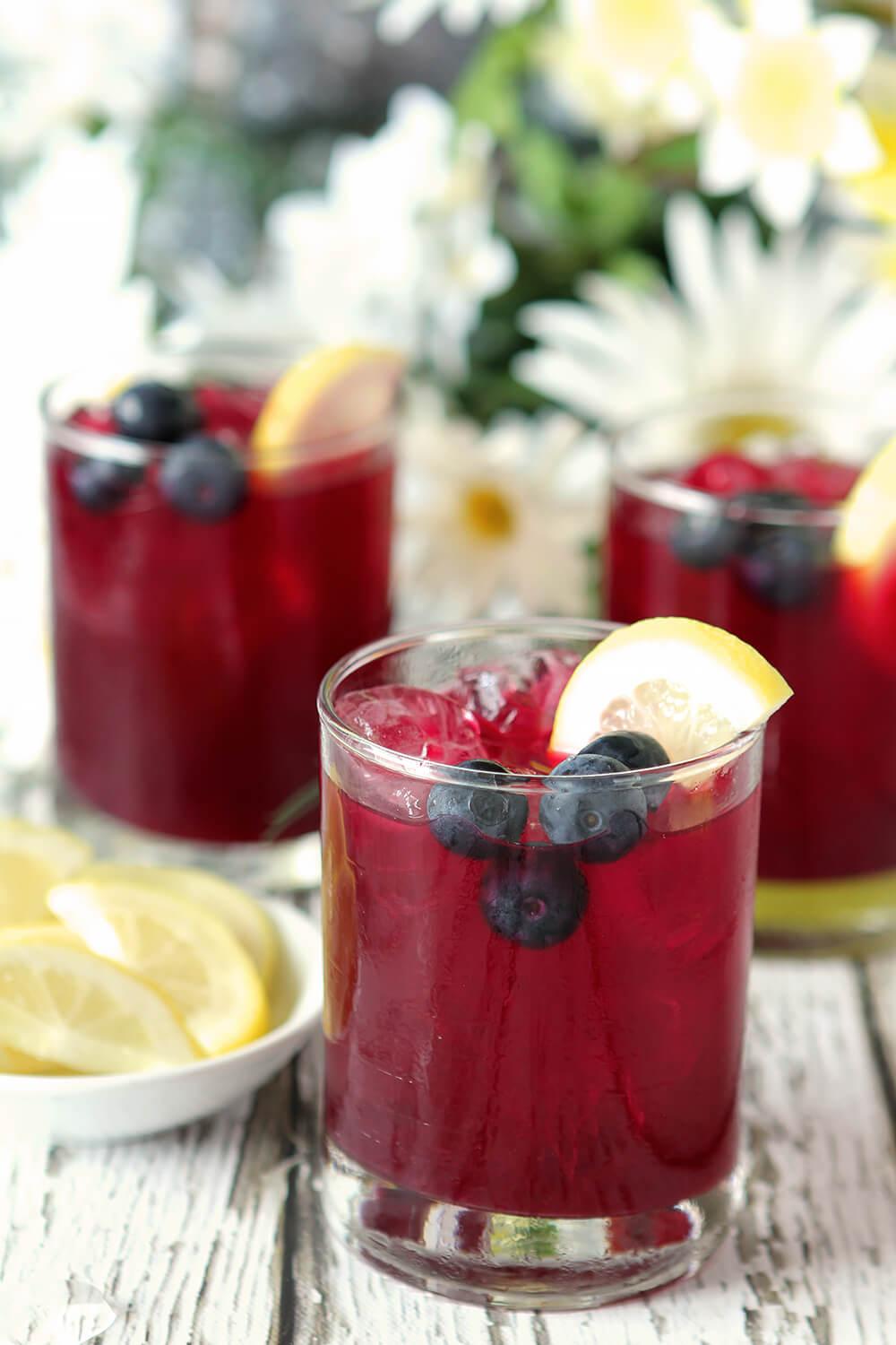 Trio of glasses of Blueberry Lemonade Spritzer with a dark burgundy beverage garnished with lemon slices and blueberries