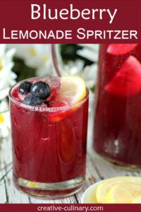 Glass of Blueberry Lemonade Spritzer with a burgundy beverage garnished with lemon slices and blueberries. Daisies in background.