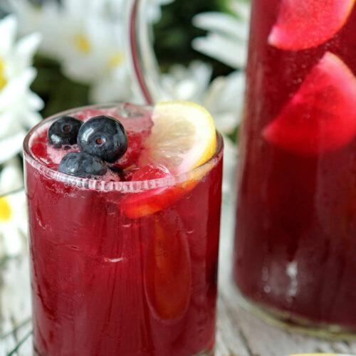 Glass of Blueberry Lemonade Spritzer with a burgundy beverage garnished with lemon slices and blueberries. Daisies in background.