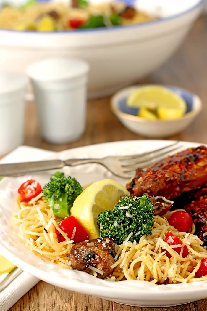 Pasta with Broccoli, Tomato, Mushrooms, and Parmesan Cheese Served with Chicken on a White Plate with Lemon Garnish