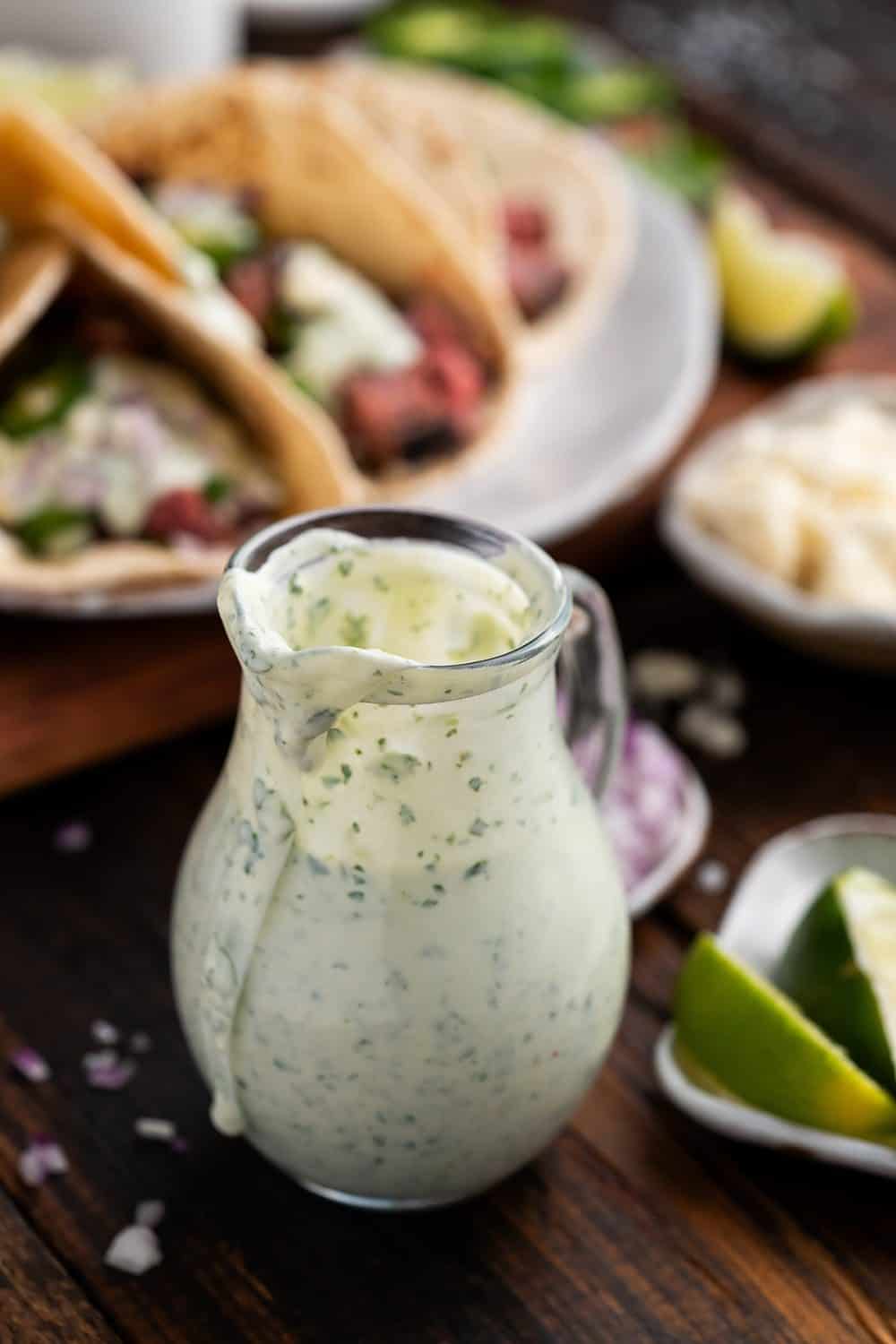 Creamy Cilantro Sauce in glass carafe and flank steak tacos in background.