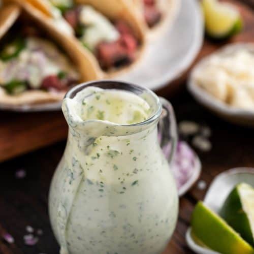 Creamy Cilantro Sauce in glass carafe and flank steak tacos in background.