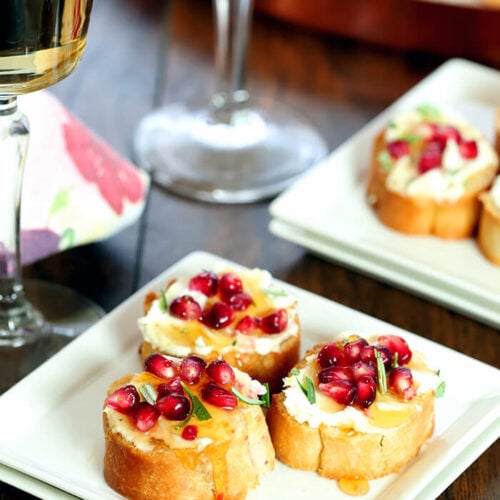 Crostini with Goat Cheese, Pomegranate Arils, and Rosemary Served on Square Plates with a Drizzle of Honey