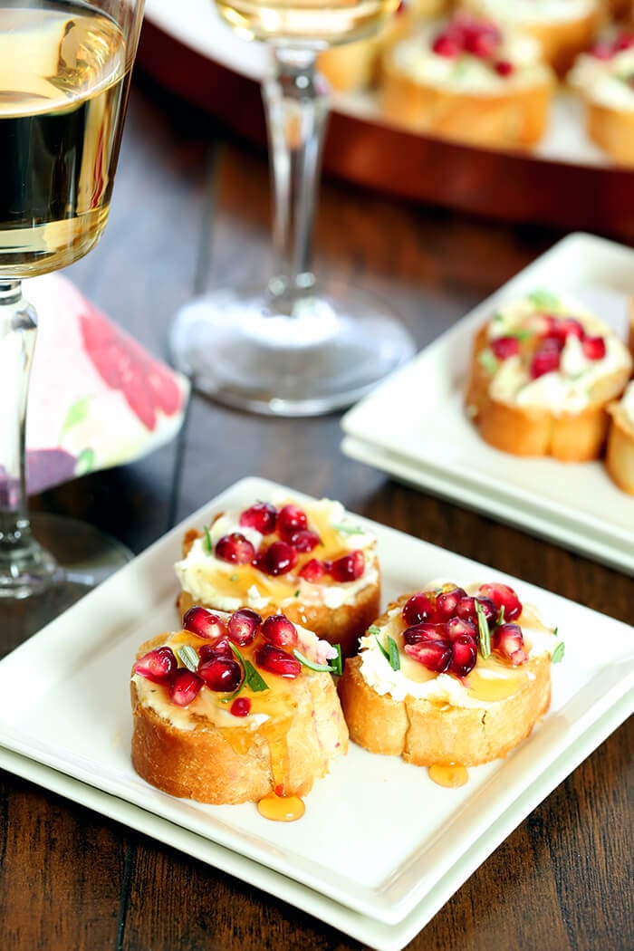 Crostini with Goat Cheese, Pomegranate Arils, and Rosemary Served on Square Plates with a Drizzle of Honey