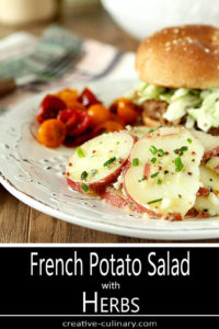 French Potato Salad with Herbs Served on a White Plate with Pork Sandwich and Cherry Tomatoes