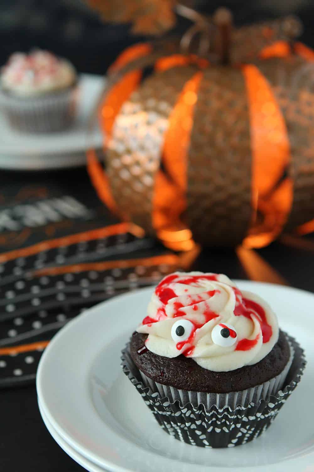 Chocolate Halloween Mummy Cupcakes with buttercream frosting and red drizzle sits on a white place on a table decorated for Halloween.