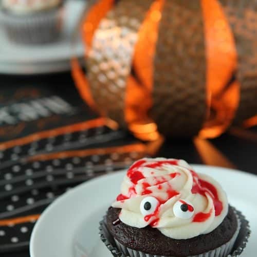 Chocolate Halloween Mummy Cupcakes with buttercream frosting and red drizzle sits on a white place on a table decorated for Halloween.