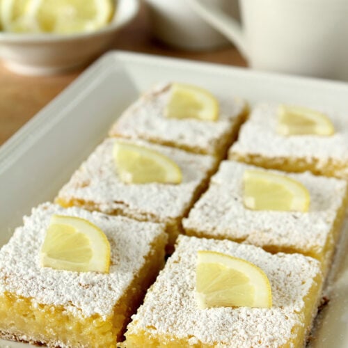 A plate of the best lemon bars cut into squares and topped with powdered sugar and a quarter slice of lemon.