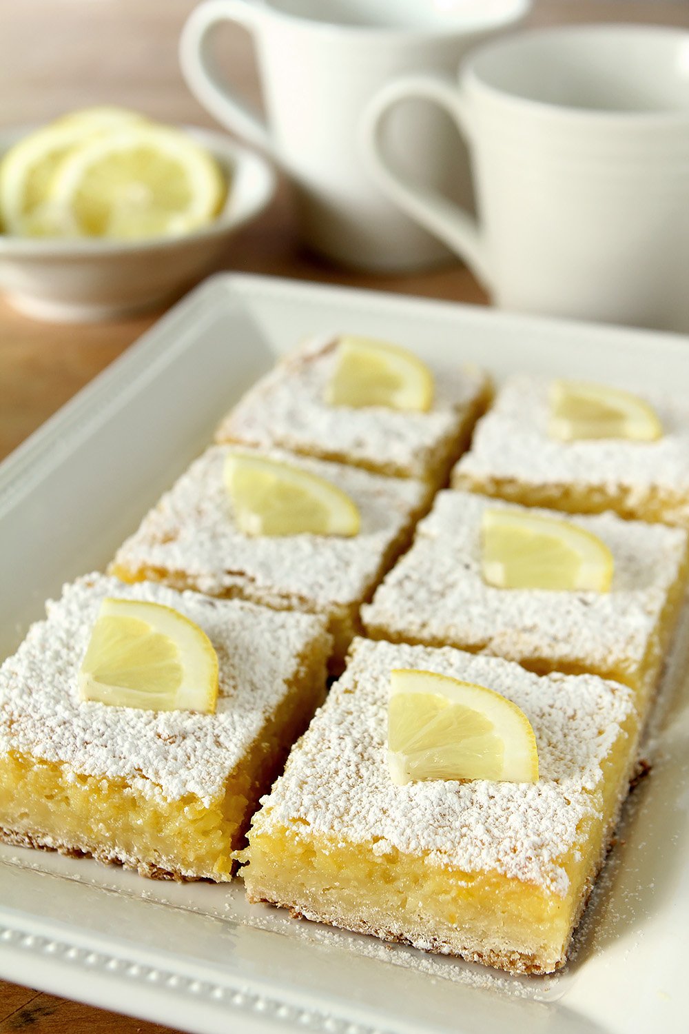 A plate of the best lemon bars cut into squares and topped with powdered sugar and a quarter slice of lemon.