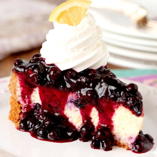 Single Slice of Lemon Blueberry Cheescake with a Blueberry Topping Served on a White Plate with Whipped Cream Topping