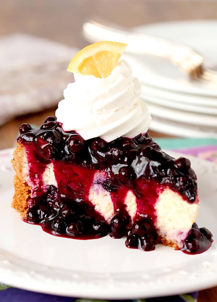 Single Slice of Lemon Blueberry Cheescake with a Blueberry Topping Served on a White Plate with Whipped Cream Topping