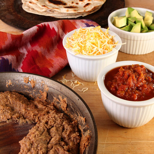 Homemade Garlic Refried Beans in a Cast Iron Skillet surrounded by bowls of Condiments