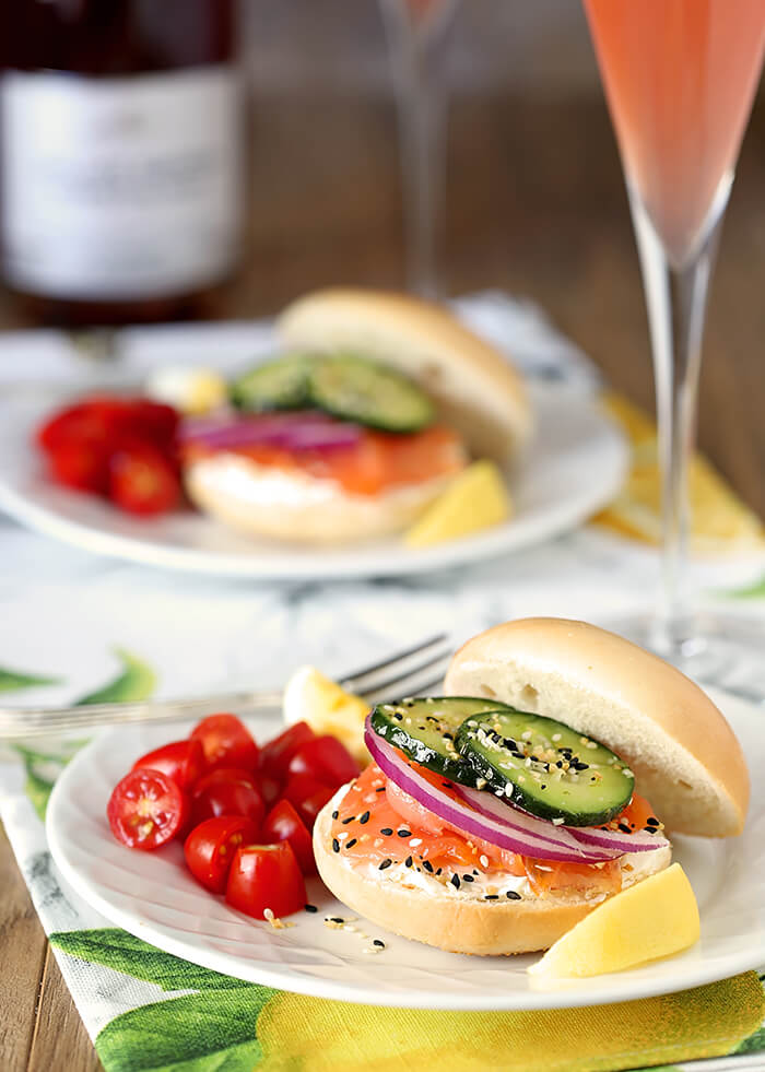 Table Setting with Bagels Stacked with Salmon, Cream Cheese, Red Onion and Cucumber on White Plates with Glasses of Pink Champagne